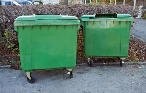 Two large green plastic refuse bins with black wheels are positioned side by side on a paved surface, likely in an outdoor urban or residential area. The bin on the left is slightly taller and has a flat, ribbed lid with a smooth surface. The bin on the right is shorter, with a similar ribbed lid, and both are situated against a background of low, leafless bushes and a paved area, suggesting late autumn or winter. The bins are designed for waste collection and are associated with rubbish removal services in Hackbridge, London postal district SW17. The environment is clean, with fallen leaves scattered on the ground, and the bins are aligned for easy access, typical of commercial or residential waste disposal points. These waste containers are representative of the types used by Commercial Waste Removal Hackbridge for waste collection and disposal in the local area, supporting waste management services in the town.