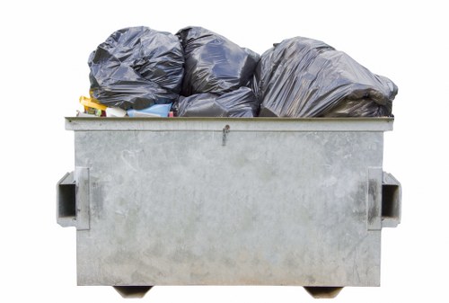 A large, industrial-style metal skip with a galvanized finish, positioned outdoors on a paved surface, is filled with black refuse bags made of thick plastic, some of which are slightly crumpled and stacked irregularly. The skip's rectangular shape features reinforced edges and side handles visible on the left and right sides. The environment suggests an urban or suburban setting, possibly near a commercial property or service area, with no other objects or background details visible. The presence of rubbish bags indicates the skip is used for waste disposal, consistent with services offered by Commercial Waste Removal Hackbridge in the local postcode area, providing rubbish removal solutions. The scene is evenly lit, emphasizing the metallic texture of the skip and the dark matte finish of the plastic bags, reinforcing the professional and neutral tone.