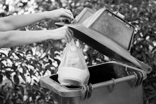 A person's hands are shown lifting the lid of a large, overfilled black wheelie bin situated outdoors on a paved or concrete surface, with leafy bushes or trees in the background. The bin is filled with various household waste, including a transparent plastic bag containing light-colored ceramic or plastic bowls. The bin lid, made of hard plastic, is partly open, revealing the contents inside. The scene suggests waste collection or rubbish disposal activity typical for commercial or residential waste management in the Hackbridge area. The image emphasizes the practicality of rubbish removal, with a focus on the type of waste that might be handled by companies like Commercial Waste Removal Hackbridge, operating within local boroughs or districts such as Sutton or nearby areas of south London.