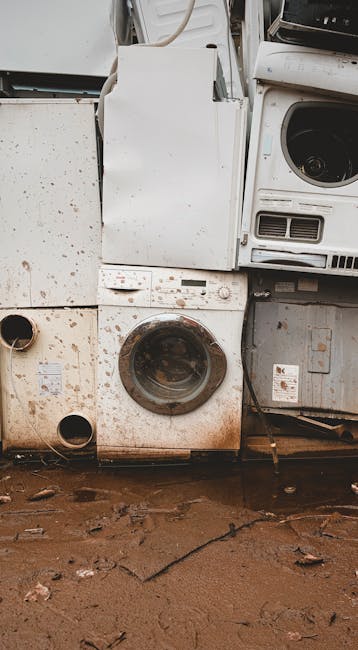 A collection of discarded household appliances stacked against a wall, including a white washing machine with visible dirt marks and rust around the door surround, a white dryer with signs of weatheri